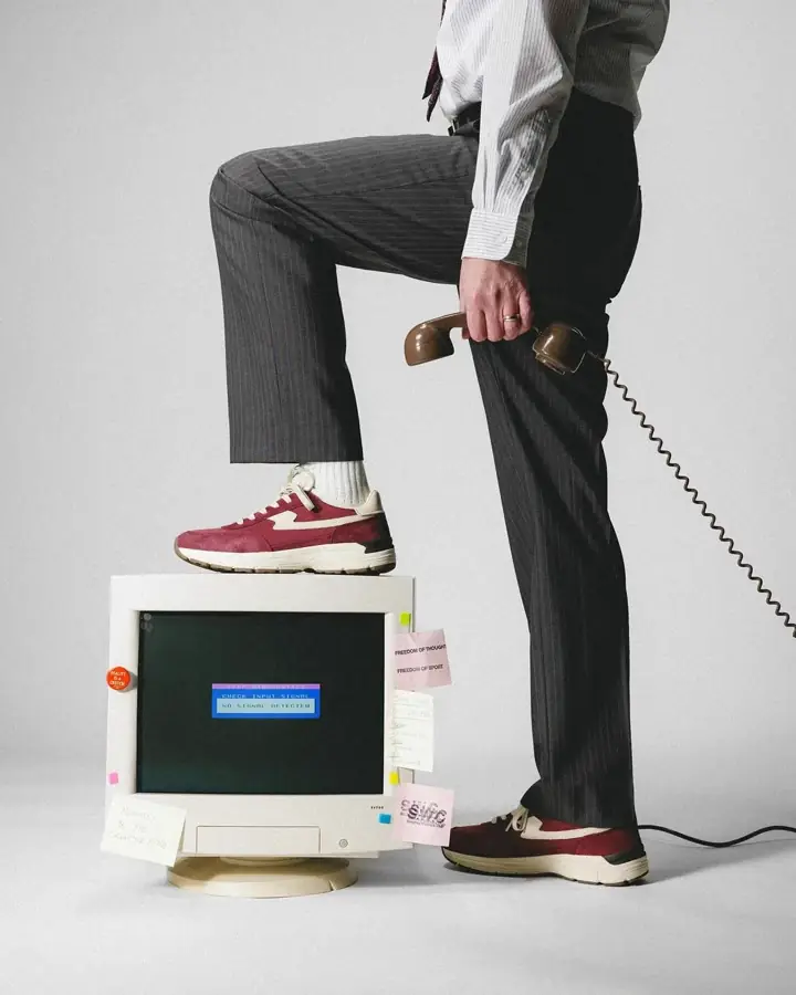 Persona trajeada con un pie apoyado sobre un monitor CRT que muestra NO SIGNAL DETECTED, sosteniendo un telefono retro